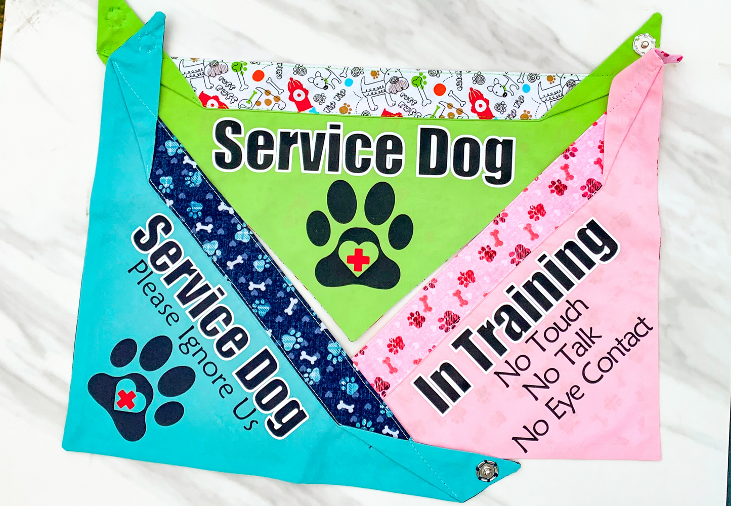 3 bandanas are shown. A green bandana with the plain paw design and the old fire hydrant and bones fabric for contrast (will be new fabric now), a blue bandana with ignore us paw design and navy paws and bone fabric, and a pink bandana with no talk design and pink bones and paws fabric for contrast.