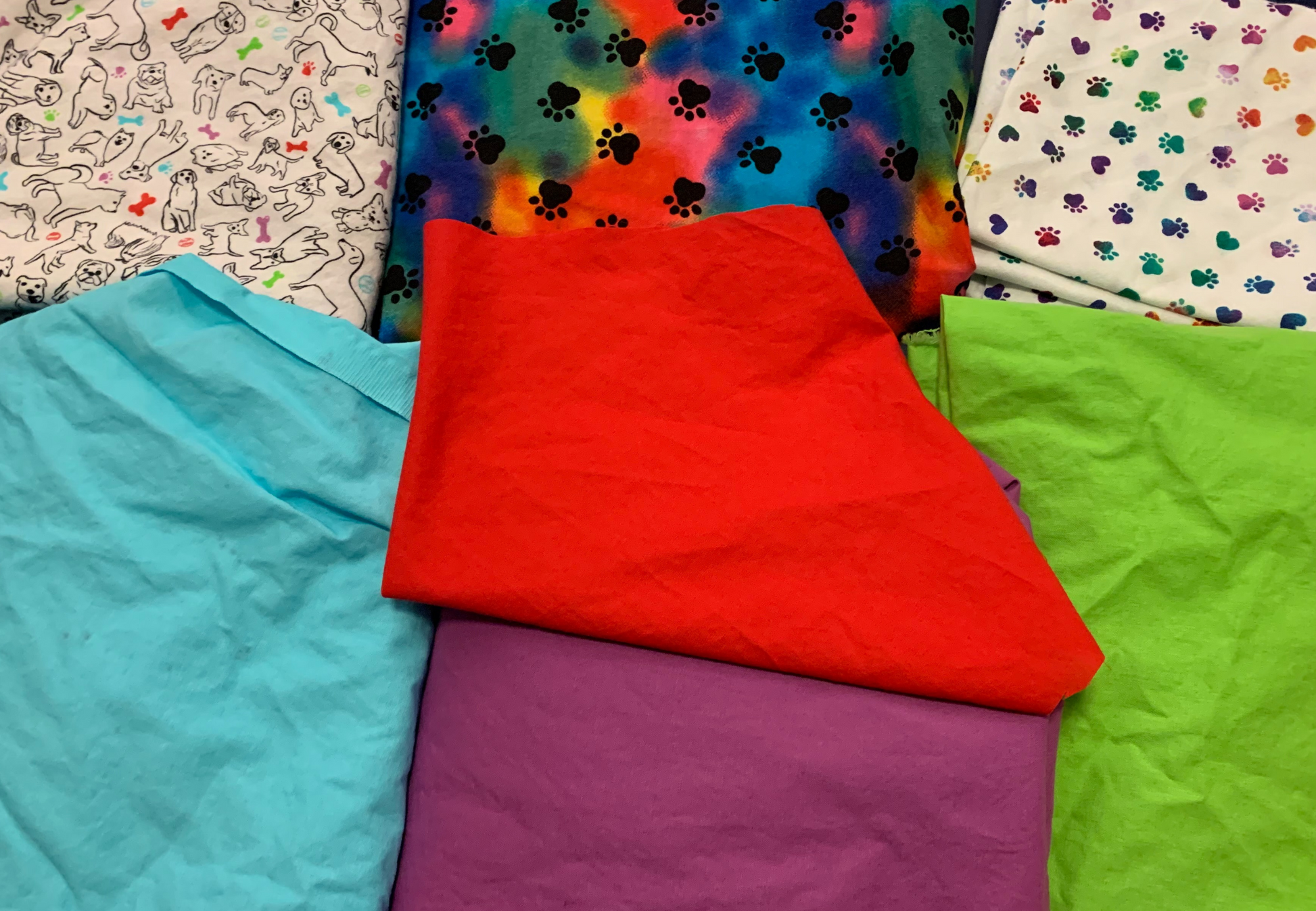 The "New" fabric is in the upper left it has a white background with different breeds of dogs drawn in black and bones, paws, and tennis balls in the colors aqua, red, purple, and green the same shades as the 4 solid color fabrics below. The upper middle fabric is Black Rainbow Paws it has a blended rainbow background and medium sized black paw prints. The fabric on the upper right is called White Rainbow Paws it has a white background and small gradient rainbow paws.