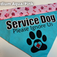 A Bandana is shown on a white surface, a label says Medium Aqua Pink. The main part of this bandana is a bright aqua color and the contrast at the top is pink paws and bones. Black text with a white outline says service dog, smaller black text below says Please Ignore Us. A black paw print below has a heart cutout in the center of the pad with a red plus in the center