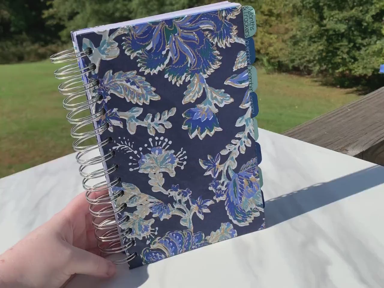 A video shows a blue and gold floral covered spiral bound book being held up, it has light and dark blue tabs that say month & trackers, then 6 months in order, and notes. An interior page is shown with days of the week, dates, what i did today, body outlines, what i ate, meds and symptom trackers and more. A grid page is shown. The top one has high medium low and is grid like, the bottom has one area per day of a month to check off. A calendar page is shown.
