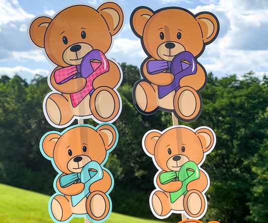 4 stickers are shown on sticks. The stickers are brown teddy bears holding an awareness ribbon with a matching colored bow tie. They are very cute. 2 stickers have white borders, 1 has a black border to blend in with the outline of the bears, and 1 sticker has a bright turquoise colored outline that is the same color of the awareness ribbon. 