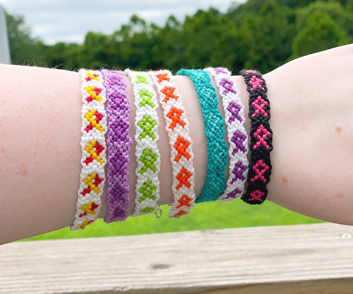 7 Bracelets are shown on a wrist. They are friendship style bracelets of knotted cotton with an awareness ribbon design. 4 of the bracelets have a white background and the ribbon colors and lime green, orange, purple, and a yellow red combo. One bracelet has a purple ribbon with a matching lighter purple background. Another has an teal ribbon with darker matching background. The last one has a black background and pink ribbon. 
