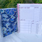 2 spiral bound books are standing up on a marble surface. The smaller one is closed and has a blue and gold floral cover. The other is open showing the interior pages. It has alternating light and dark pink tabs that are blank. The page has several sections and the titles have a pink shadow.