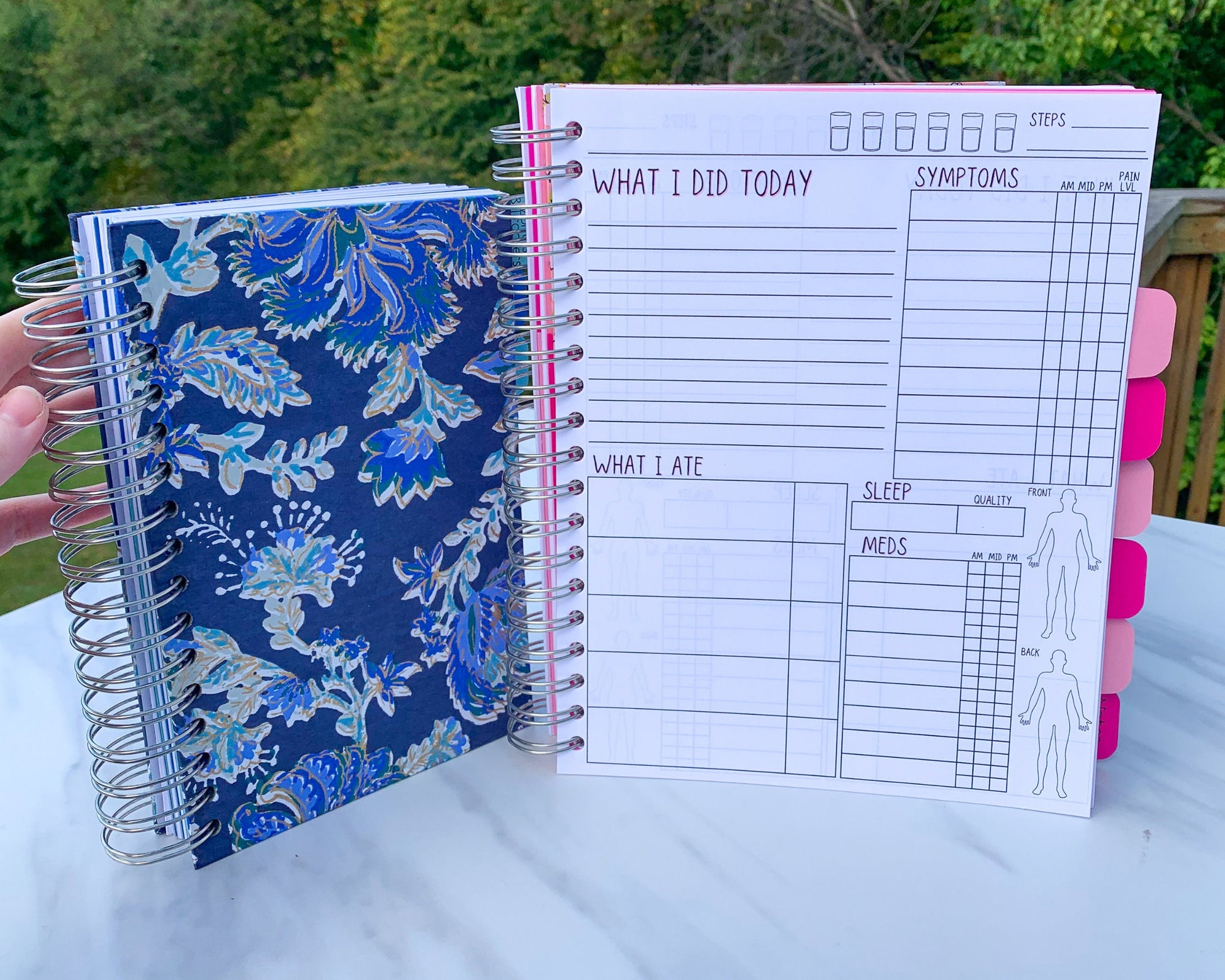 2 spiral bound books are standing up on a marble surface. The smaller one is closed and has a blue and gold floral cover. The other is open showing the interior pages. It has alternating light and dark pink tabs that are blank. The page has several sections and the titles have a pink shadow.