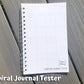 The spiral tester is shown on grey wood planks. It is closed and you can see the spoon culture logo on the front as well as the side of the coils. Text says spiral journal tester.