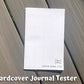 The hardcover tester is shown closed on grey wood planks. You can see the spoon culture logo on the bottom right and text that says symptom journal tester. On the left edge of the booklet you can see the back side of 3 staples. Text on the page says Hardcover Journal Tester.