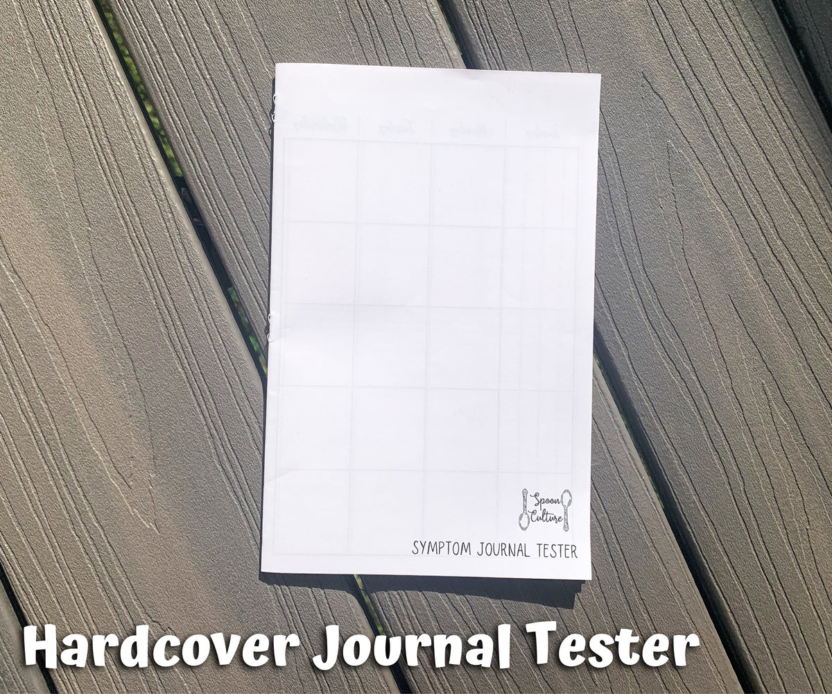 The hardcover tester is shown closed on grey wood planks. You can see the spoon culture logo on the bottom right and text that says symptom journal tester. On the left edge of the booklet you can see the back side of 3 staples. Text on the page says Hardcover Journal Tester.