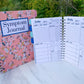 2 spiral bound books are standing up on a marble surface. The larger one is closed and has a pink floral cover with a blue outlined label that says Symptom Journal. The other is open showing the interior pages. It has alternating light and dark blue tabs that are labeled with months. The page has several section and the titles have a blue shadow. 