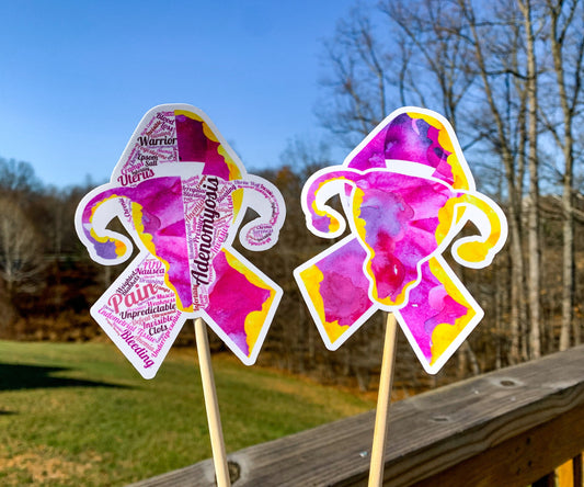 The word cloud and watercolor designs are shown on 2 sticks. Both ribbons feature a magenta/pink color and irregular yellow border on the edges. The design an awareness ribbon with a uterus in the center. The word cloud design on the left is split in half with a word cloud on the left half of the ribbon and right half of the uterus. 