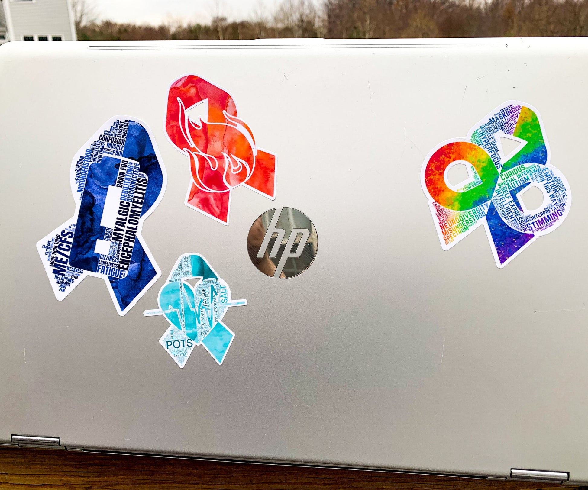 A silver laptop is shown with a 4 1/2 inch word cloud MECFS ribbon compared to a 3 1/2 inch watercolor CRPS and word cloud autism sticker, and a 2 1/2 inch word cloud POTS sticker.