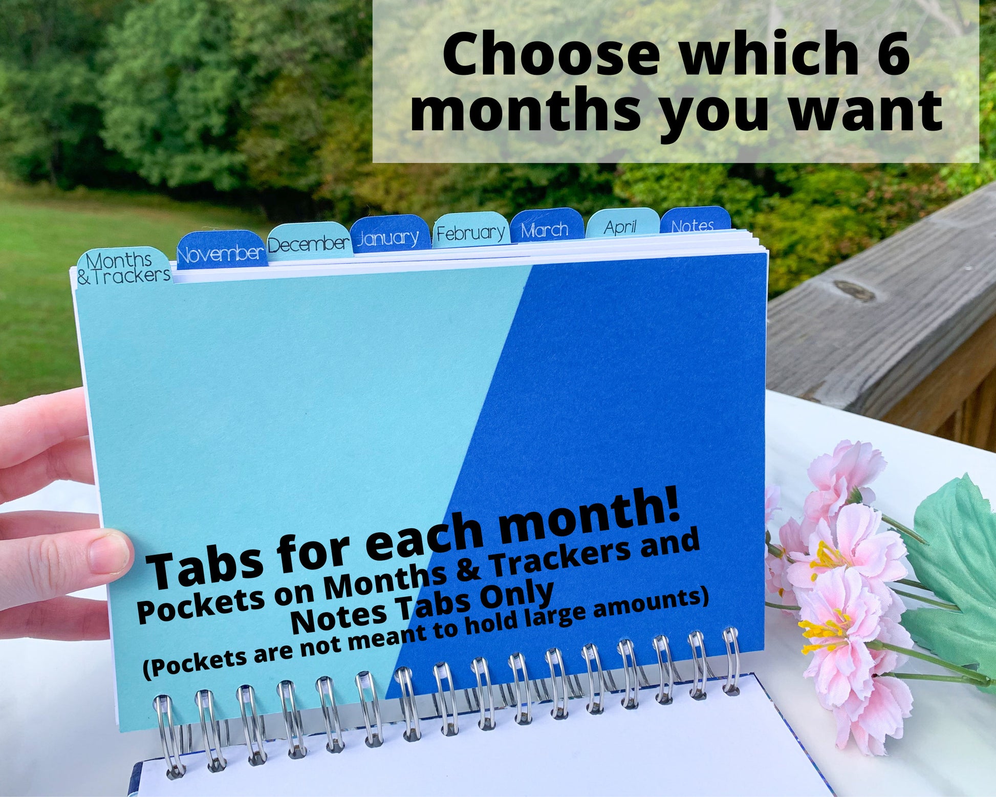 The book is being held up without the cover to show the tabs. The first tab is labeled Months and trackers, it is light blue with a darker blue angled pocket. The other tabs are November to April and Notes. The tabs alternate between light blue and dark blue, with white writing on the darker tabs. A note says choose which 6 months you want. Another note says Tabs for each month, pocket on months and tracker, and notes tabs only. Pockets are not meant to hold large amounts.