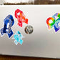 A silver laptop is shown with a 4 1/2 inch word cloud MECFS ribbon compared to a 3 1/2 inch watercolor CRPS and word cloud autism ribbon and a 2 1/2 inch word cloud POTS sticker.