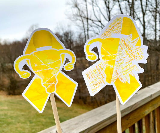 The word cloud and watercolor designs are shown on 2 sticks. Both ribbons are yellow with a shape in the center, the left watercolor one is a full uterus with barbed wire and the word cloud is half uterus and barbed wire and half sunflower. The word cloud design on the left is split in half with a word cloud on the left half of the ribbon and right sunflower half. The right watercolor ribbon is all color with a white border around the uterus for definition.