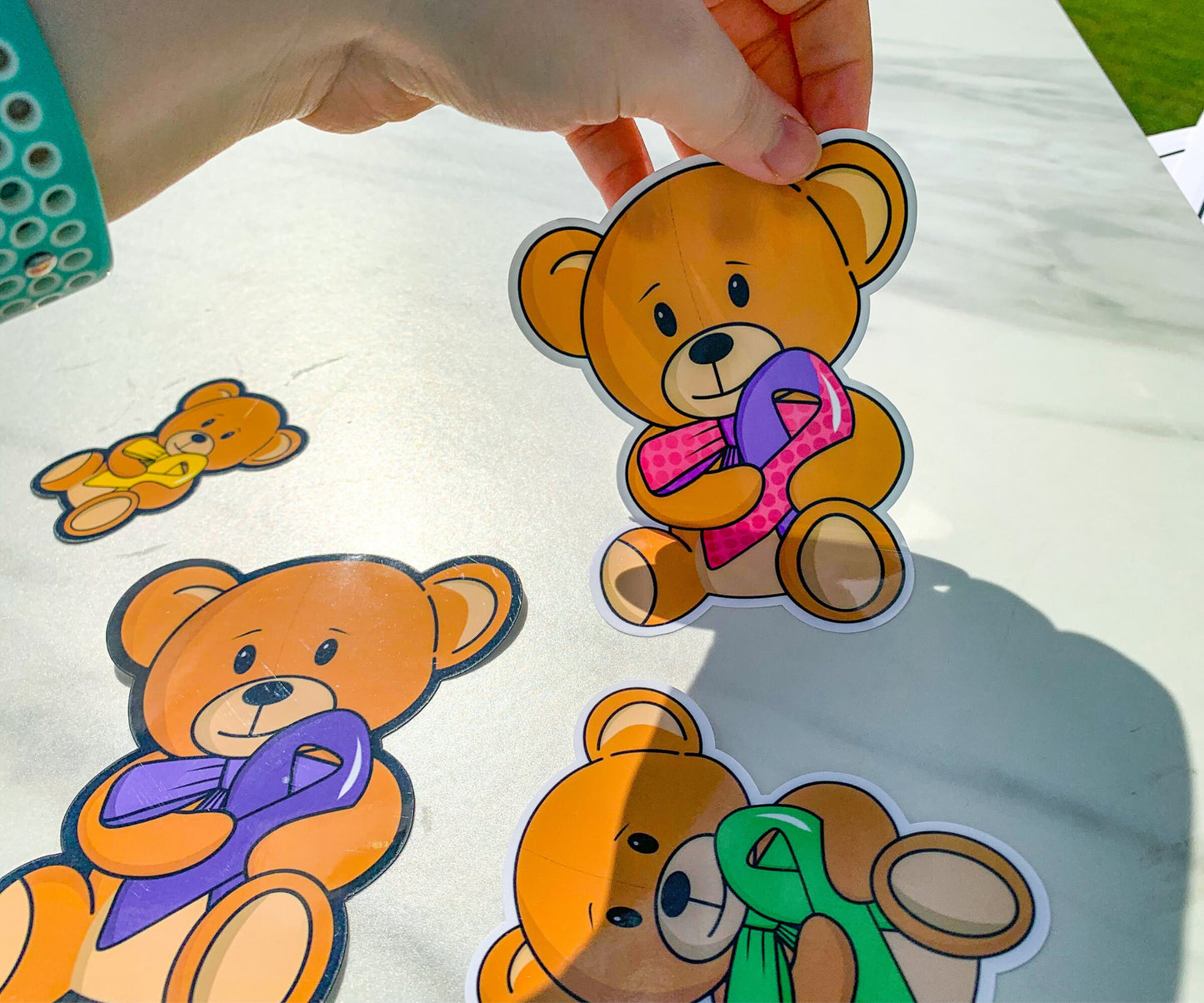 A hand is peeling up a teddy bear sticker from a marble surface. The bear has a mixed ribbon with one purple side and one pink polka dot side. The bow tie has the same dot pattern and same colors. Purple, green, and yellow ribbon bears are on the marble surface. This is to demostrate that the stickers can peel off.