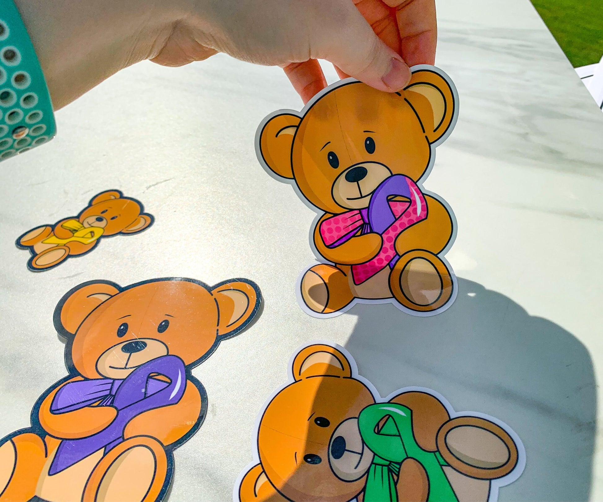 A hand is peeling up a teddy bear sticker from a marble surface. The bear has a mixed ribbon with one purple side and one pink polka dot side. The bow tie has the same dot pattern and same colors. Purple, green, and yellow ribbon bears are on the marble surface. This is to demostrate that the stickers can peel off.