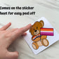A bear holding a lesbian pride flag with a matching bow tie is shown being peeled off sticker paper. The sticker comes on the sticker sheet for easy peel off.