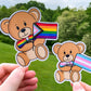2 stickers are shown being held up. The stickers are brown teddy bears holding pride flags with a matching colored bow tie. They are very cute. The stickers have a white outline of the teddy bear.